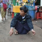 Autoostas ļaudis. Kungs svārkos un ar cigareti, tupēdams gaida savu autobusu.